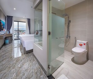 Habitación de hotel moderna con ducha de vidrio, balcón con vista a la ciudad, cama bien hecha y baño con inodoro y ducha.