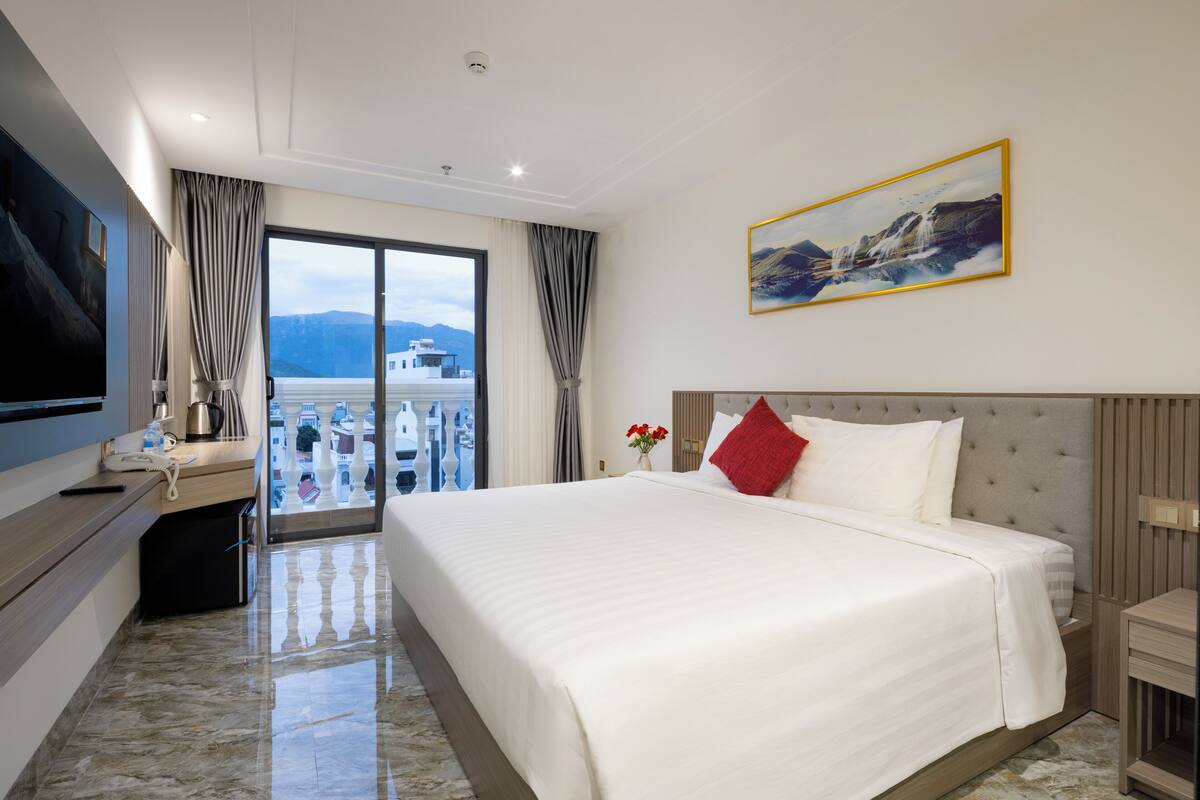 A hotel room with a large bed, a flat-screen TV, a desk with a telephone, and a view of a cityscape through a balcony.