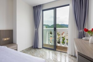 Habitación de hotel con cama, mesita de noche, escritorio con teléfono y florero, y balcón con vista a las montañas.