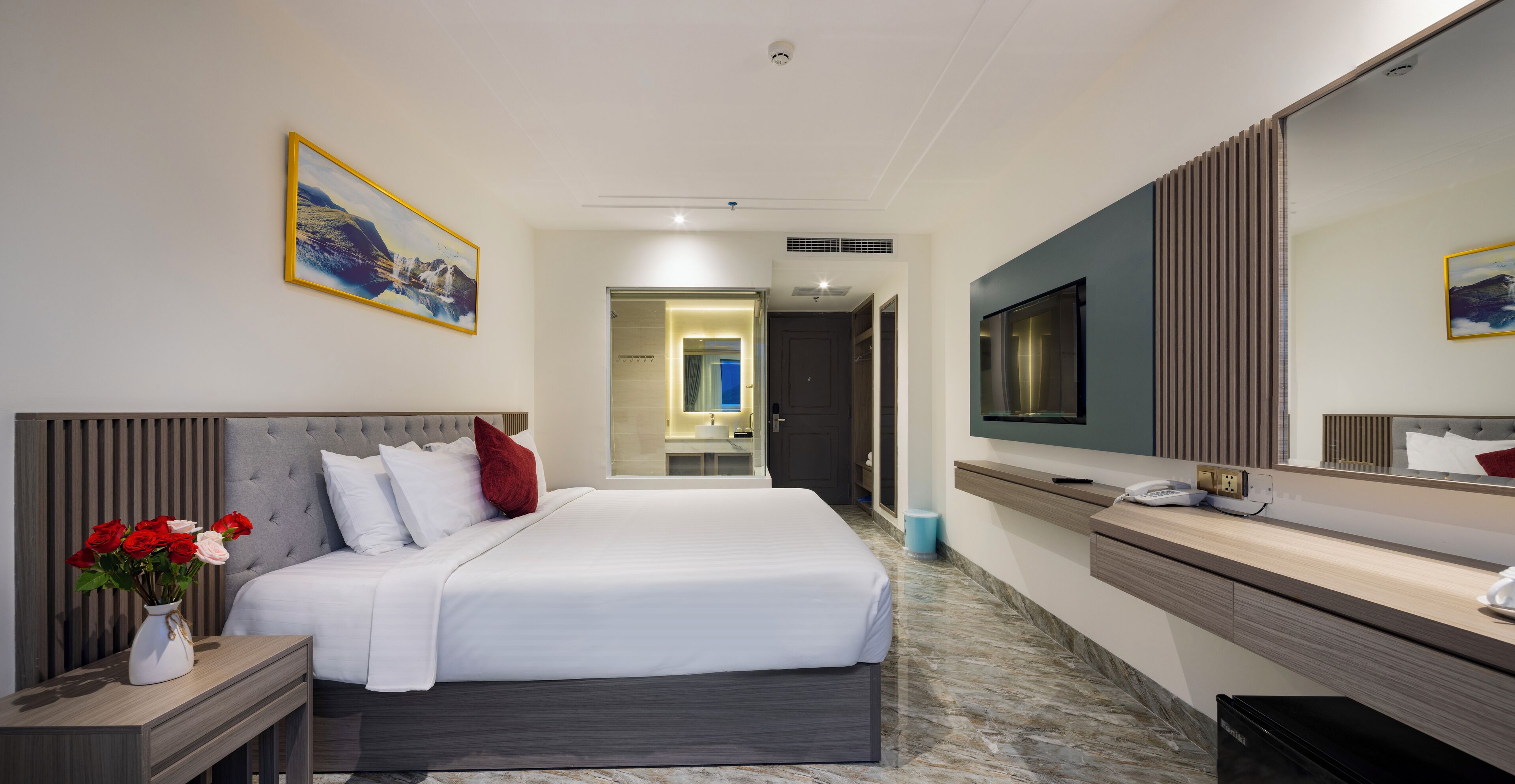 Ein modernes Hotelzimmer mit einem großen Bett, einem Nachttisch mit Blumenvase, einem großen Spiegel, einem Fernseher und einem durch eine offene Tür einsehbaren Badezimmer.
