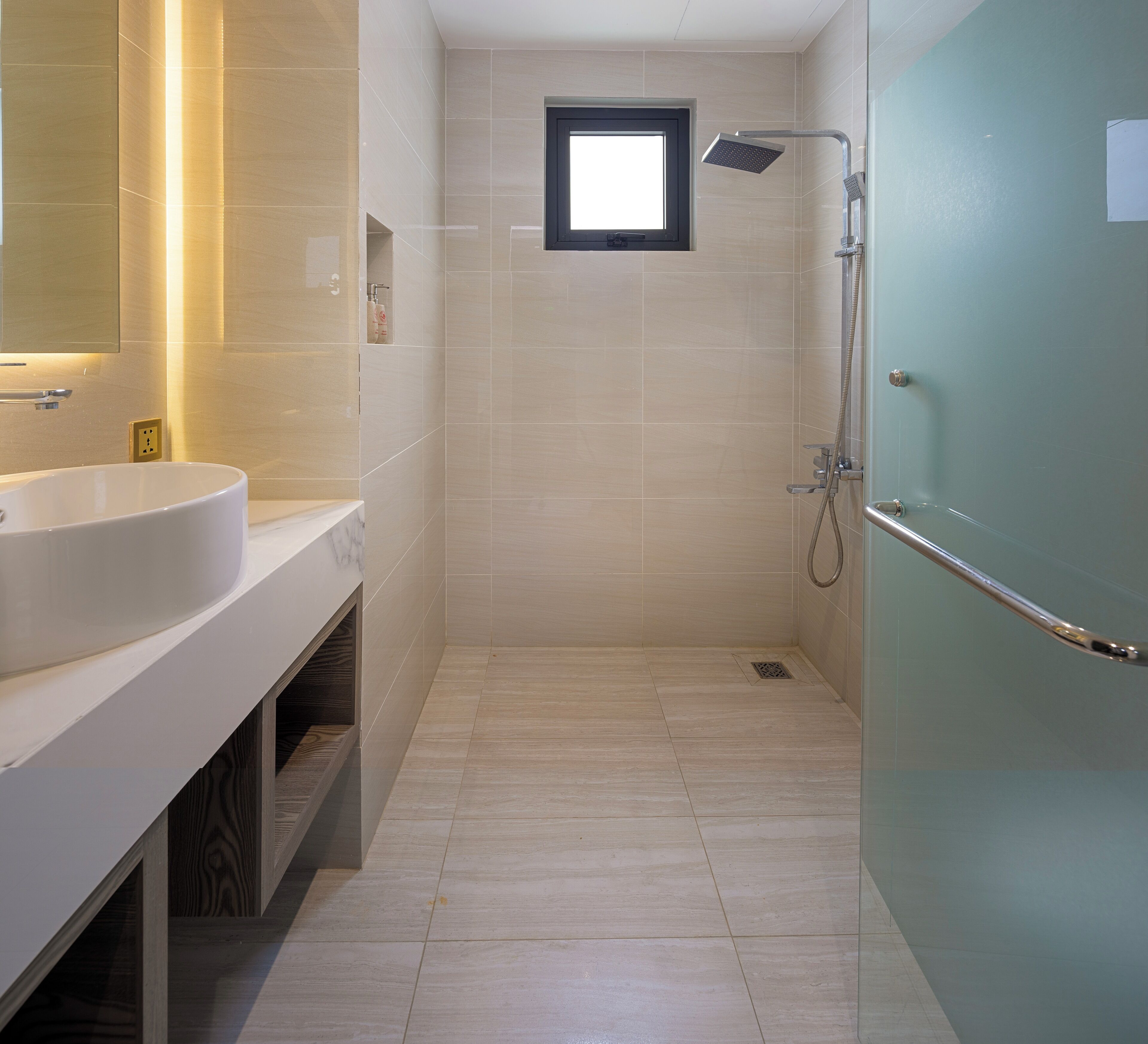 A modern bathroom with a large sink, a shower with a handheld showerhead, and a small window.