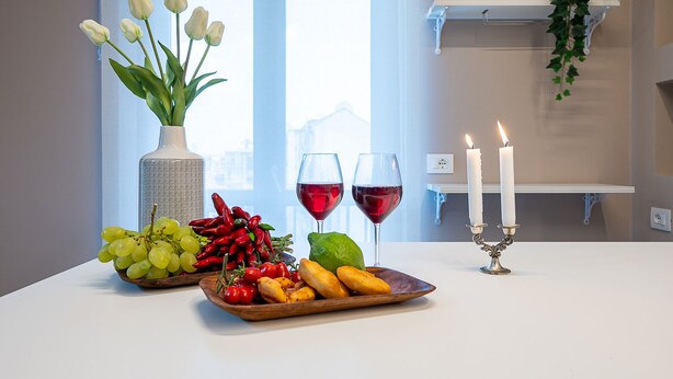 A table with a vase of white tulips, a bowl of grapes, red chili peppers, a lime, and a plate of fried plantains. There are two glasses of red wine and two lit candles on a white table. - [2min InalpiArena] Cozy Suite + A/C + WI-FI (Torino)