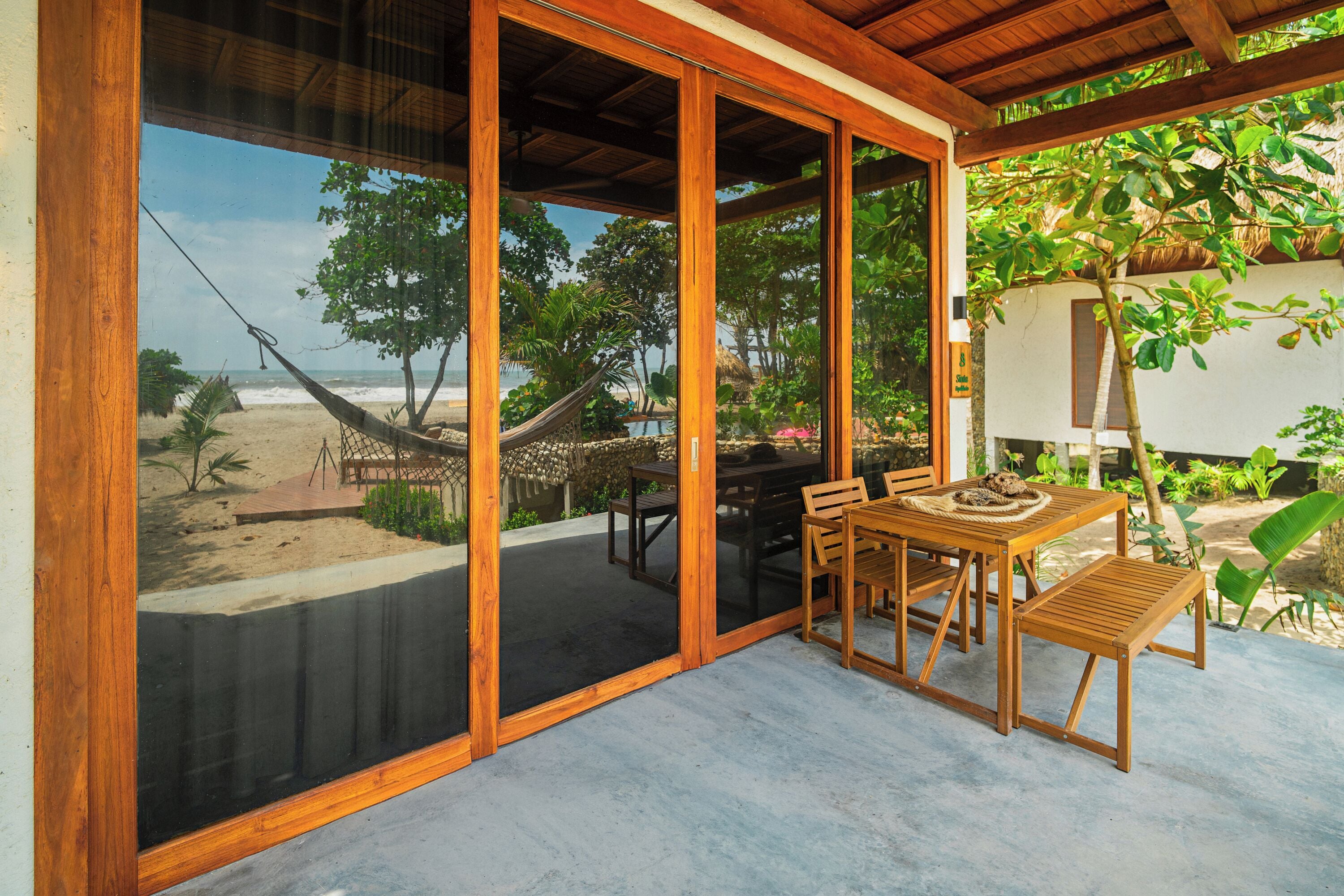 A covered outdoor seating area with a view of the beach, wooden furniture, and a hammock.