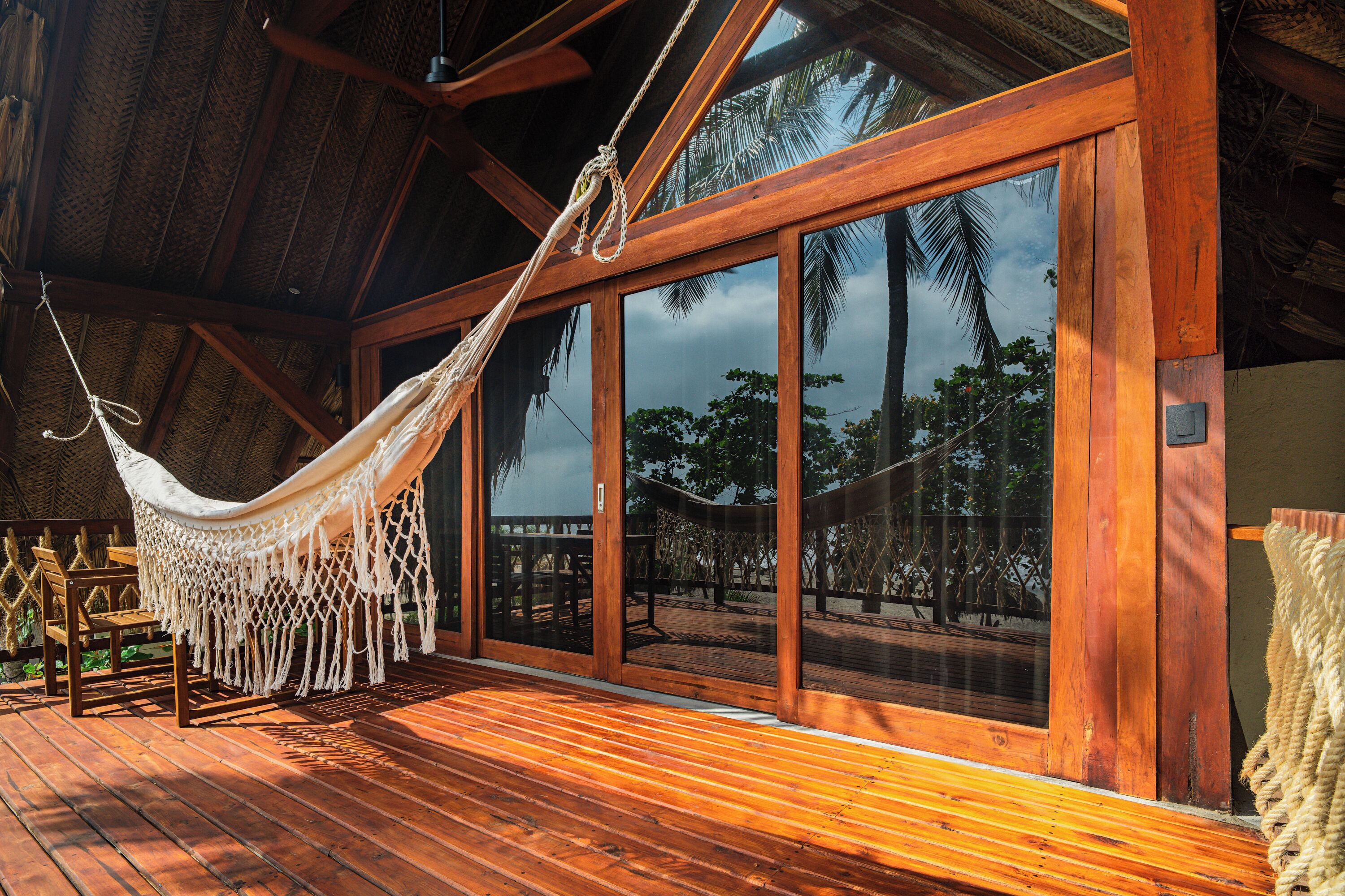 A wooden deck with a hammock and a wooden chair, large glass doors, and a view of palm trees.
