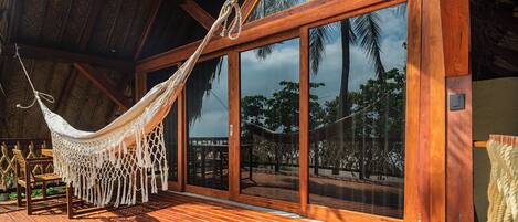 A wooden deck with a hammock and a wooden chair, large glass doors, and a view of palm trees.