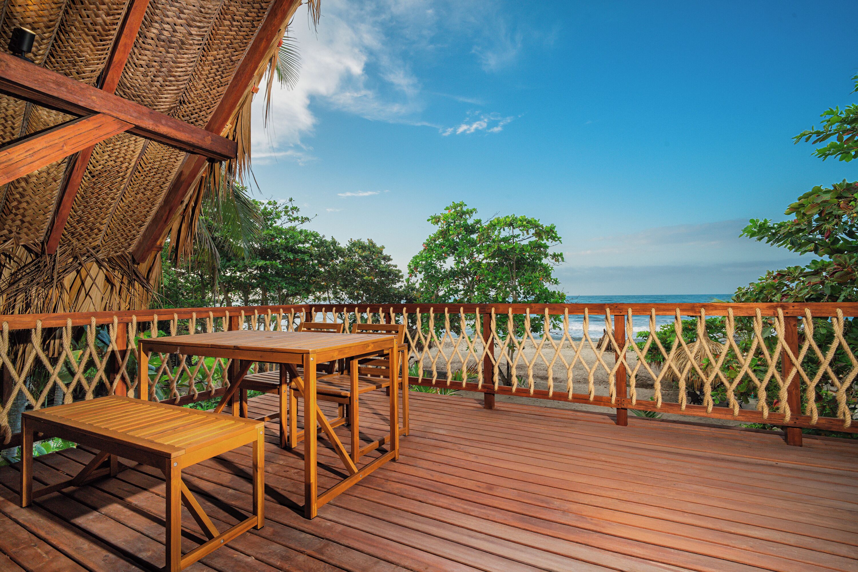 A wooden deck with a table and bench, overlooking a beach with a thatched roof structure and a wooden fence.