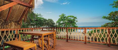 A wooden deck with a table and bench, overlooking a beach with a thatched roof structure and a wooden fence.