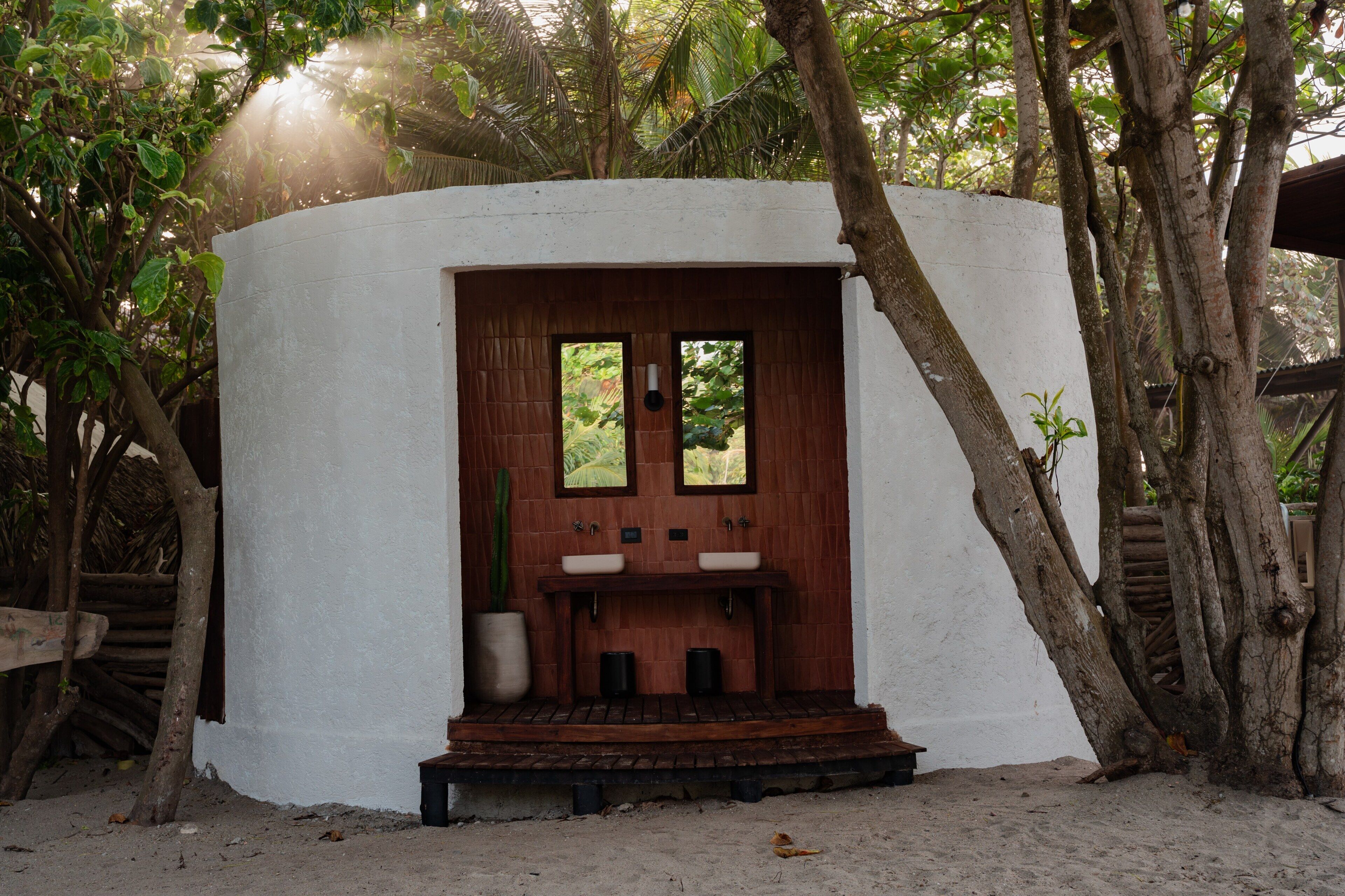 Une petite construction blanche, composée d’une seule pièce, avec une porte en bois, deux fenêtres et un évier.