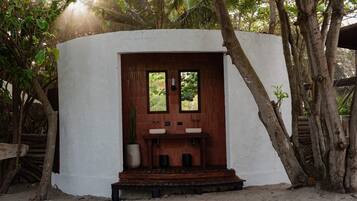 Une petite construction blanche, composée d’une seule pièce, avec une porte en bois, deux fenêtres et un évier.