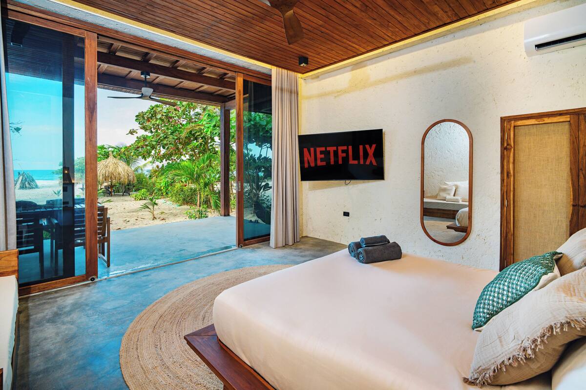 A bedroom with a large bed, a TV mounted on the wall, and a view of the beach through the sliding glass door.
