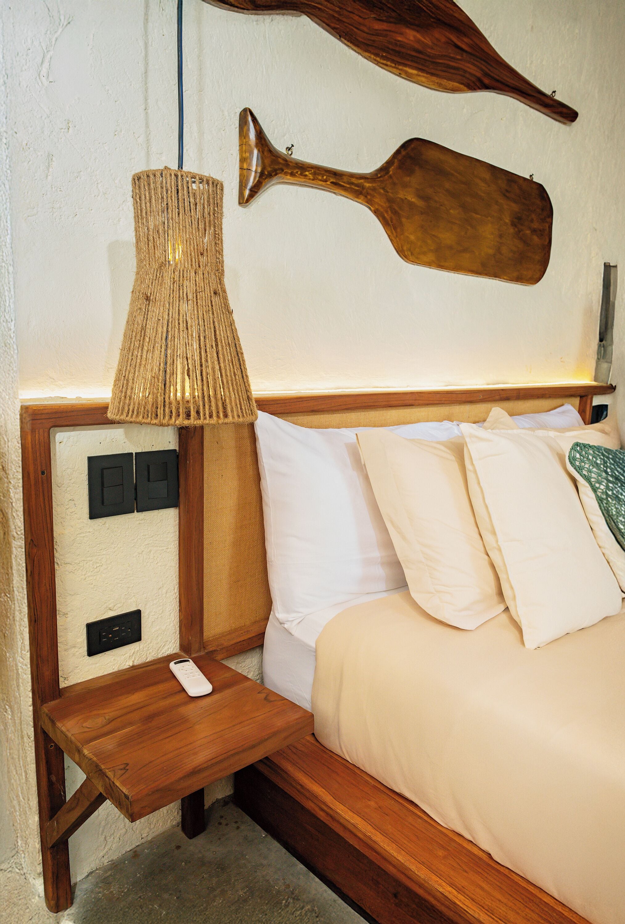 A neatly made bed with white linens, a wooden nightstand with a remote control, a woven lamp, and two wall-mounted wooden objects.
