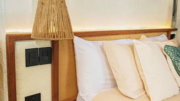 A neatly made bed with white linens, a wooden nightstand with a remote control, a woven lamp, and two wall-mounted wooden objects.