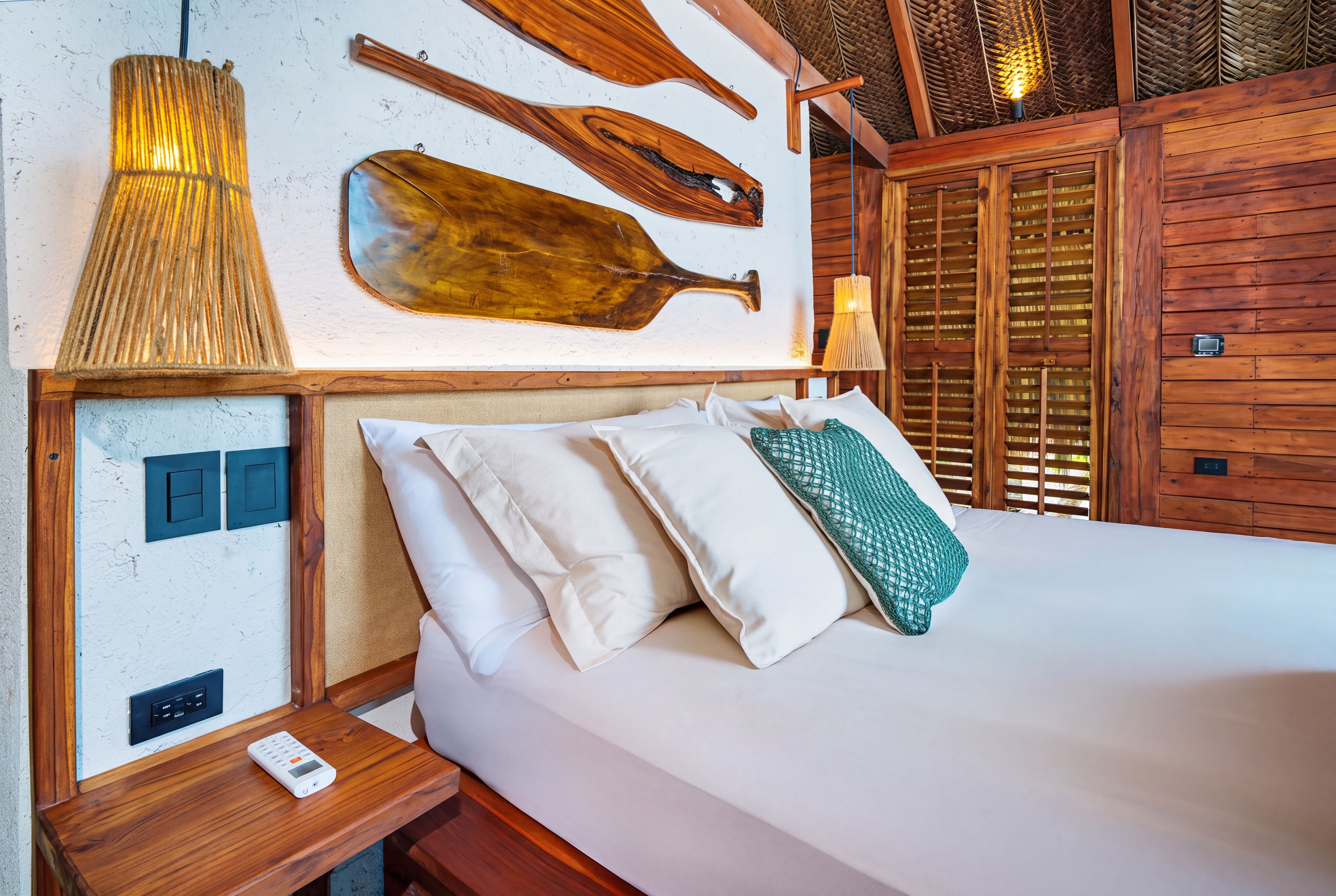 A hotel room with a wooden headboard, a bed with white linens, a nightstand with a digital thermostat, and a wall-mounted light fixture.