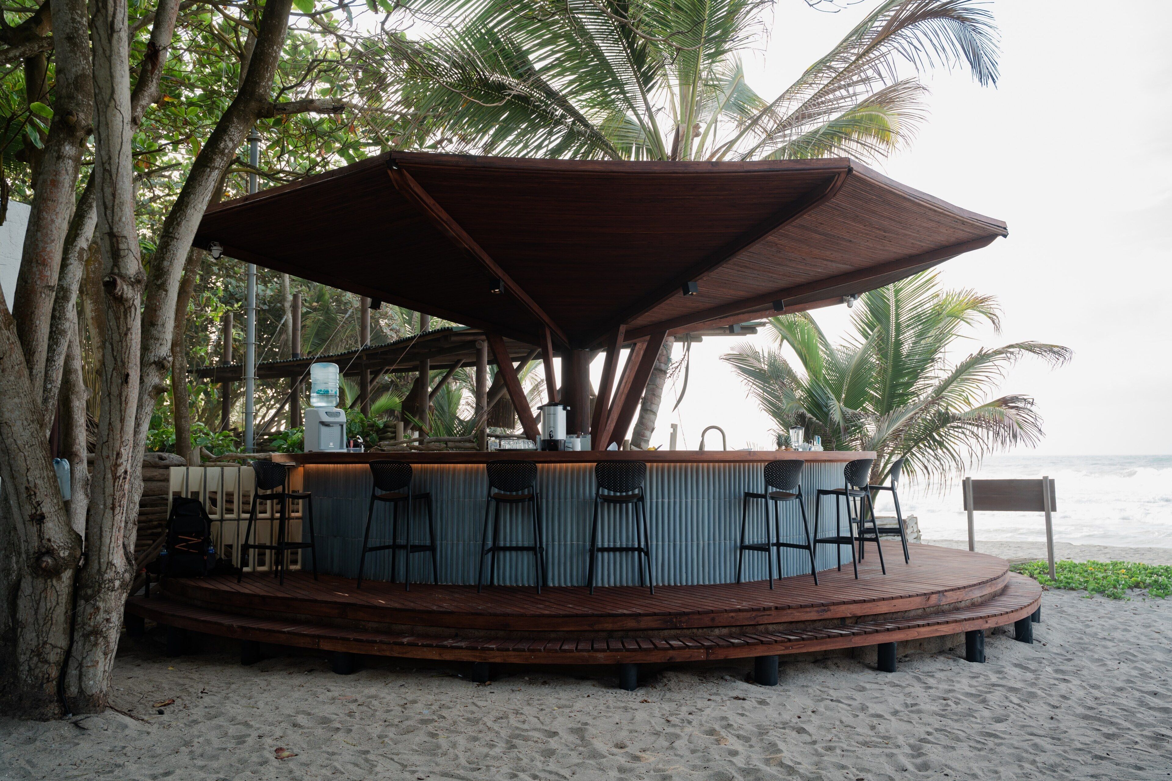Un bar junto a la playa con una terraza de madera, taburetes y una zona de estar cubierta.