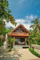 A beachfront bungalow with a thatched roof, a wooden balcony, and a hammock.
