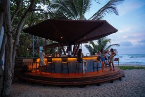 An outdoor bar area with a wooden deck, bar stools, and a large umbrella providing shade. - Casa Iwana Tayrona (Santa Marta)