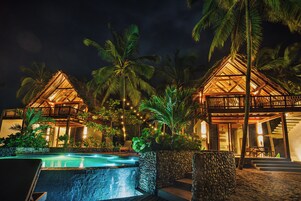 A resort with two thatched-roof villas, a swimming pool, and lush tropical vegetation.