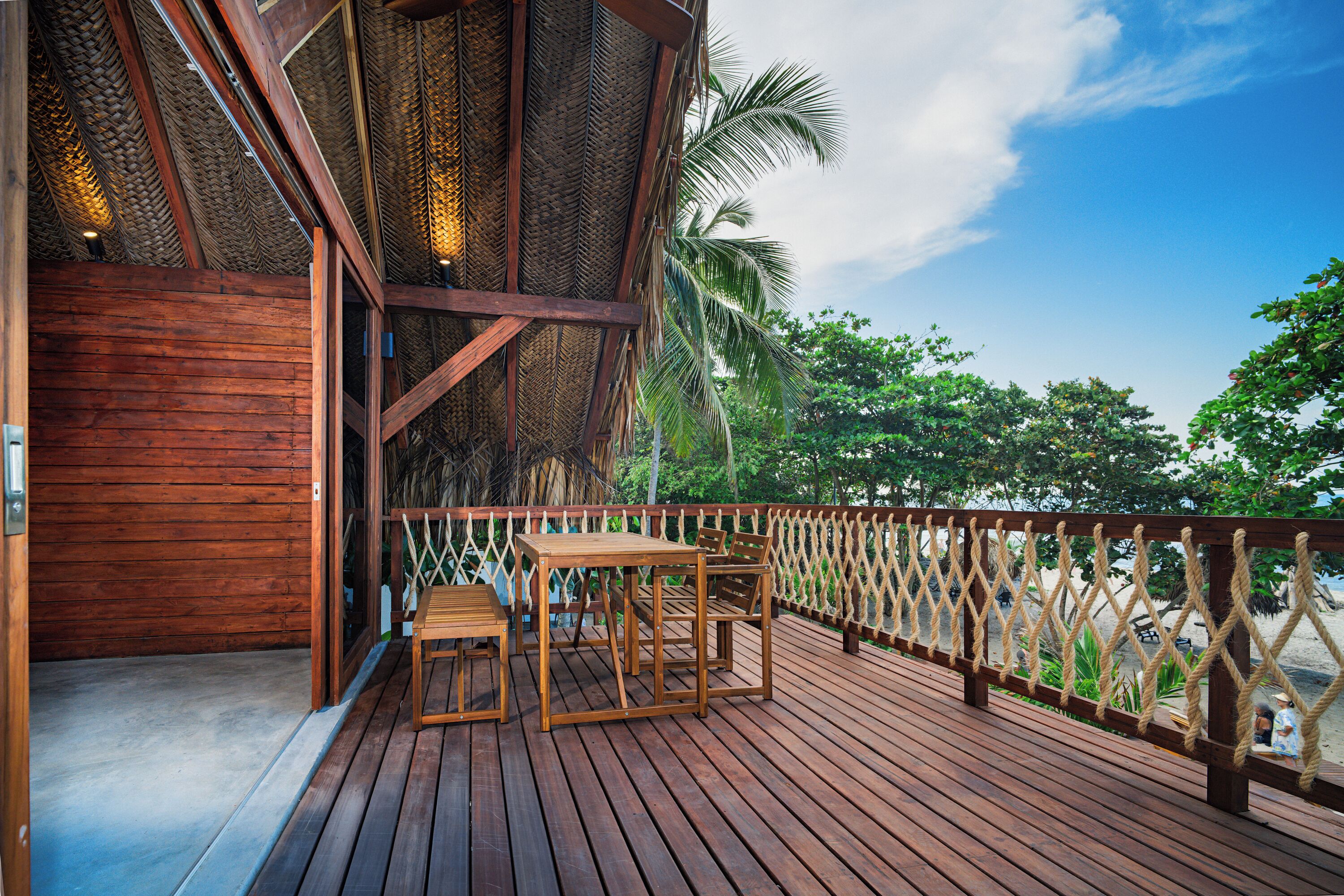 A wooden deck with a table and chairs, overlooking a beach with palm trees.