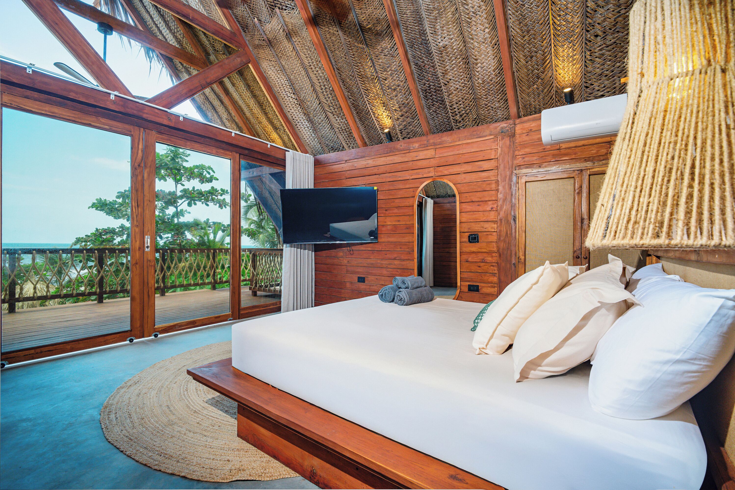 A bedroom with a large bed, a flat-screen TV, and a view of the ocean through large glass doors.