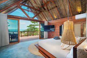 A bedroom with a large bed, a TV mounted on the wall, and a view of the ocean through a sliding glass door.