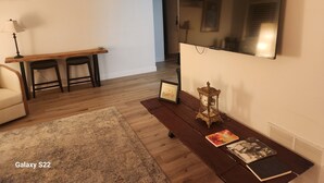 A living room with a wooden bench, a small table, a sofa, a TV mounted on the wall, and a framed picture on the floor. - Stylish Coastal Condo Retreat. Bike to Beach & Dining. Complimentary Parking (Point Pleasant)