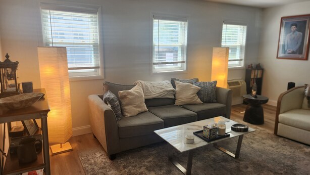 A living room with a gray sofa, a coffee table, and a lamp. There are windows with blinds, a framed picture on the wall, and a small table with a lamp. - Stylish Coastal Condo Retreat. Bike to Beach & Dining. Complimentary Parking (Point Pleasant)