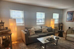 A living room with a gray sofa, a coffee table, and a lamp. There are windows with blinds, a framed picture on the wall, and a small table with a lamp.