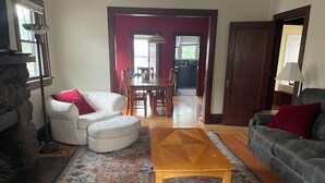 Ein Wohnzimmer mit einem weißen Sofa, einem grauen Sofa, einem Holztisch und einem gemusterten Teppich. Im Hintergrund ein Essbereich mit einer roten Wand, und durch eine offene Tür ist eine Küche zu sehen.