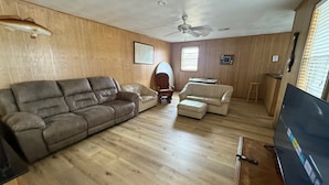 Een woonkamer met een bruine leren bank, een witte leren zithoek, een houten vloer, een plafondventilator en een televisie.