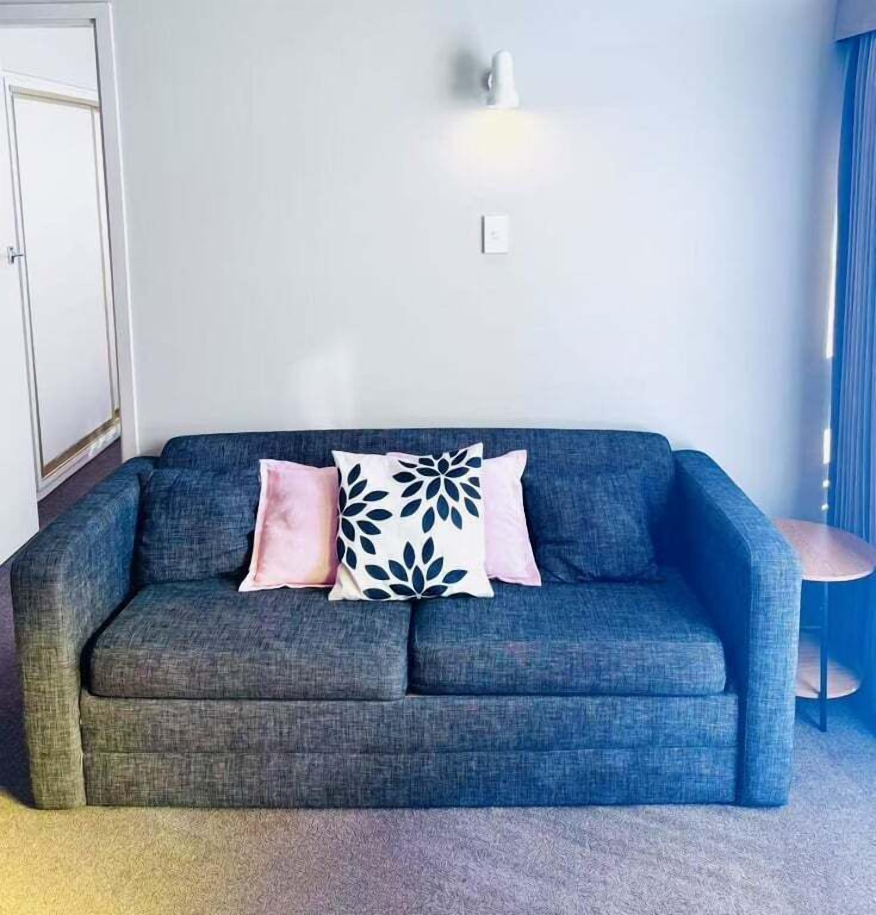 A grey sofa with two pillows, one pink and one with a leaf pattern, against a plain wall with a light switch and a small table with a lamp.