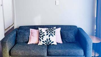 A grey sofa with two pillows, one pink and one with a leaf pattern, against a plain wall with a light switch and a small table with a lamp.