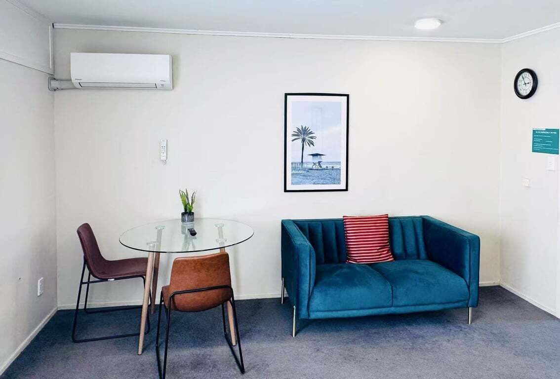 A small, modern room with a glass table and two chairs, a teal sofa, a framed picture of a beach scene, and a clock on the wall.