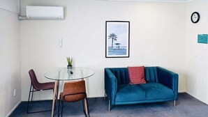 A small, modern room with a glass table and two chairs, a teal sofa, a framed picture of a beach scene, and a clock on the wall.