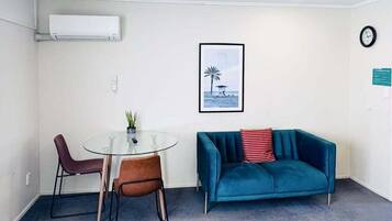 A small, modern room with a glass table and two chairs, a teal sofa, a framed picture of a beach scene, and a clock on the wall.