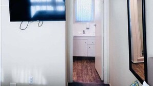 A room with a flat-screen TV mounted on the wall, a door leading to a bathroom with a sink and mirror, and a large mirror on the wall.