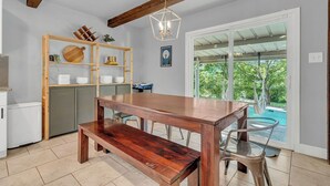 A dining area with a wooden table and bench, a metal chair, a wooden shelf with kitchen items, and a view of a swimming pool through a glass door.