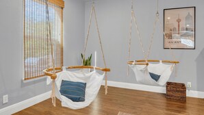A room with two hanging hammocks, a window with blinds, a framed cityscape picture, and a wooden floor.