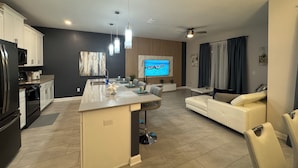 Una cocina moderna con una isla central, un comedor con sofá y un televisor que muestra una vista panorámica.