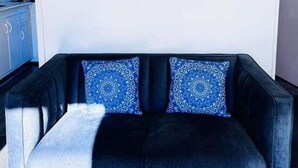 Un canapé bleu foncé agrémenté de coussins à motifs, un tableau représentant une orchidée accroché au mur, et un coin cuisine en arrière-plan.