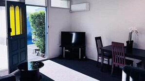 Une pièce avec un téléviseur, une table à manger avec des chaises, une plante en pot et une porte donnant sur l’extérieur.
