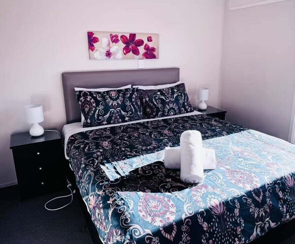 Een net opgemaakt bed met een gebedrukte sprei, twee kussens en een bedlampje op een nachtkastje.