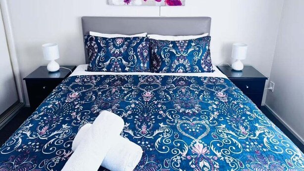 Un lit bien fait, recouvert d’une courtepointe à motifs floraux bleus et agrémenté d’oreillers assortis, avec une tête de lit grise et une œuvre d’art florale accrochée au mur au-dessus du lit.