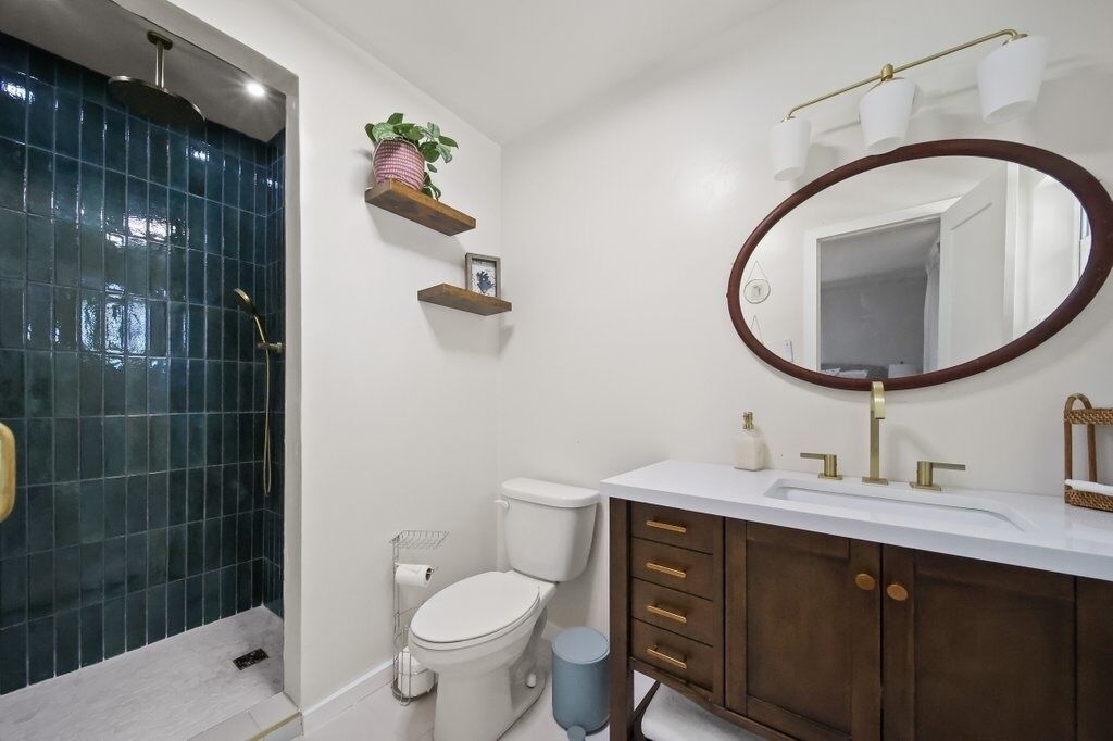 A bathroom with a shower, toilet, vanity with a mirror, and shelves with plants.