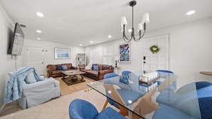 A modern living room with a glass dining table, blue chairs, a brown leather sofa, and a ceiling light fixture. - Tropical Retreat • Pool • 1.5 miles to Beach (Hobe Sound)