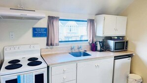 Une cuisine de taille réduite équipée d’une cuisinière, d’un four à micro-ondes, d’un évier et de placards. Un panneau indique qu’il faut laver la vaisselle.