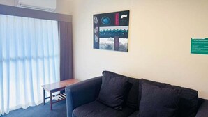 Une pièce avec un canapé de couleur sombre, une petite table en bois et une grande fenêtre ornée de rideaux légers.