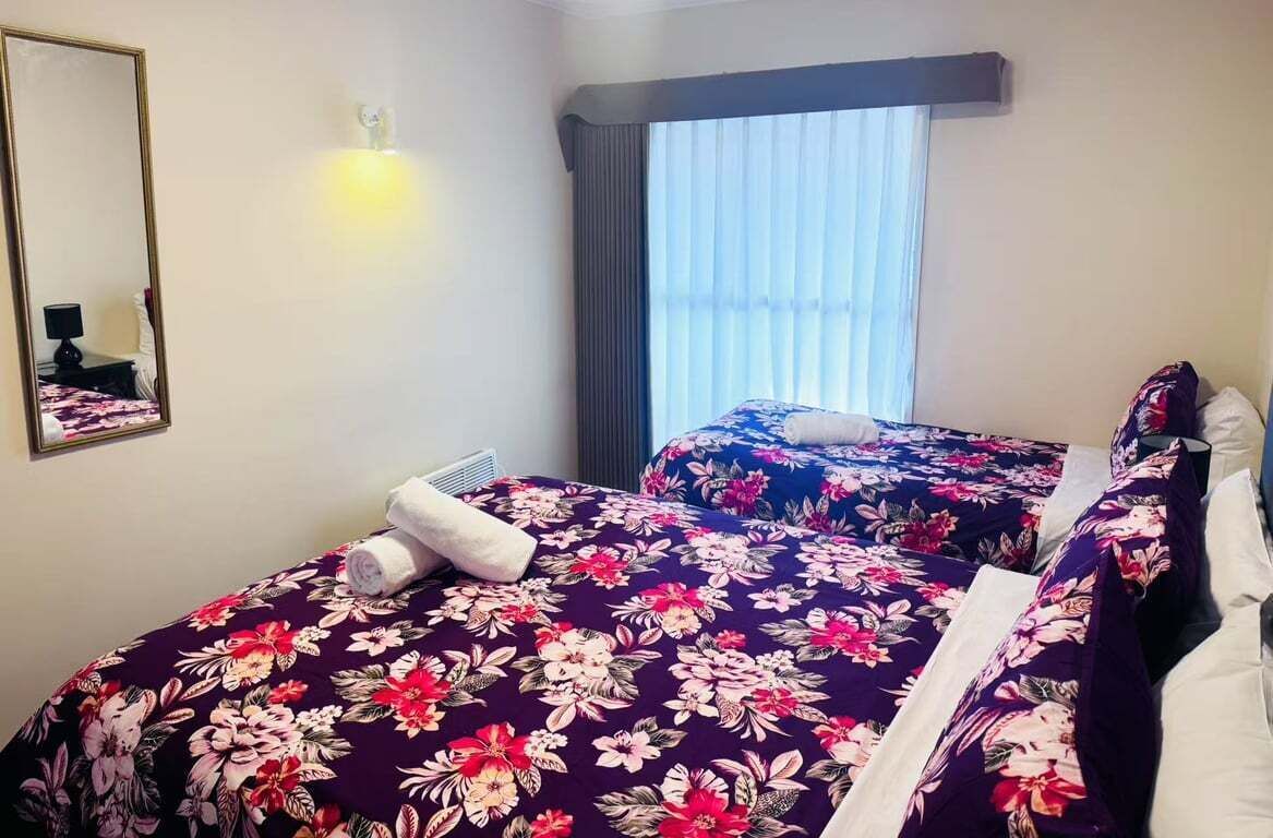 A hotel room with two beds, floral bedspreads, a mirror, and a window with curtains.