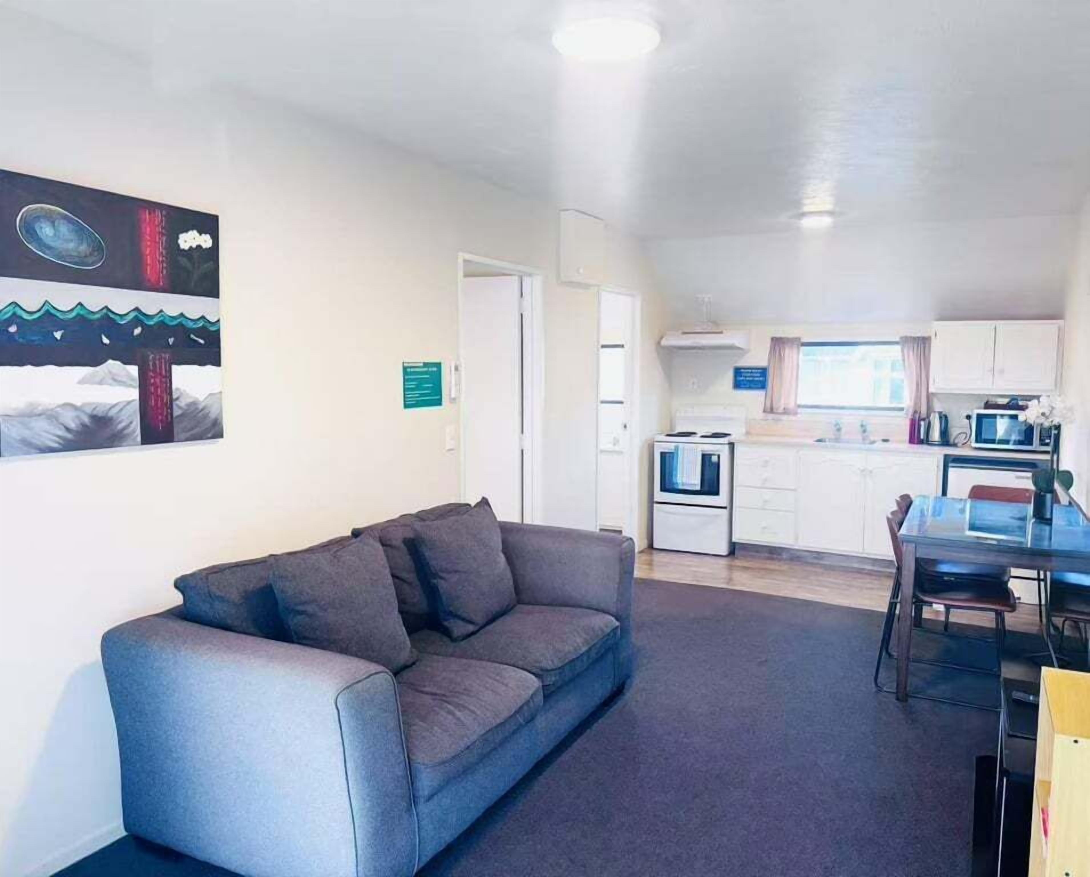 A living room with a grey sofa, a dining table with chairs, a kitchen area with a stove and microwave, and a painting on the wall.