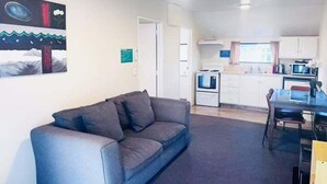 Un salon avec un canapé gris, une table à manger avec des chaises, un coin cuisine avec une cuisinière et un four à micro-ondes, et un tableau accroché au mur.
