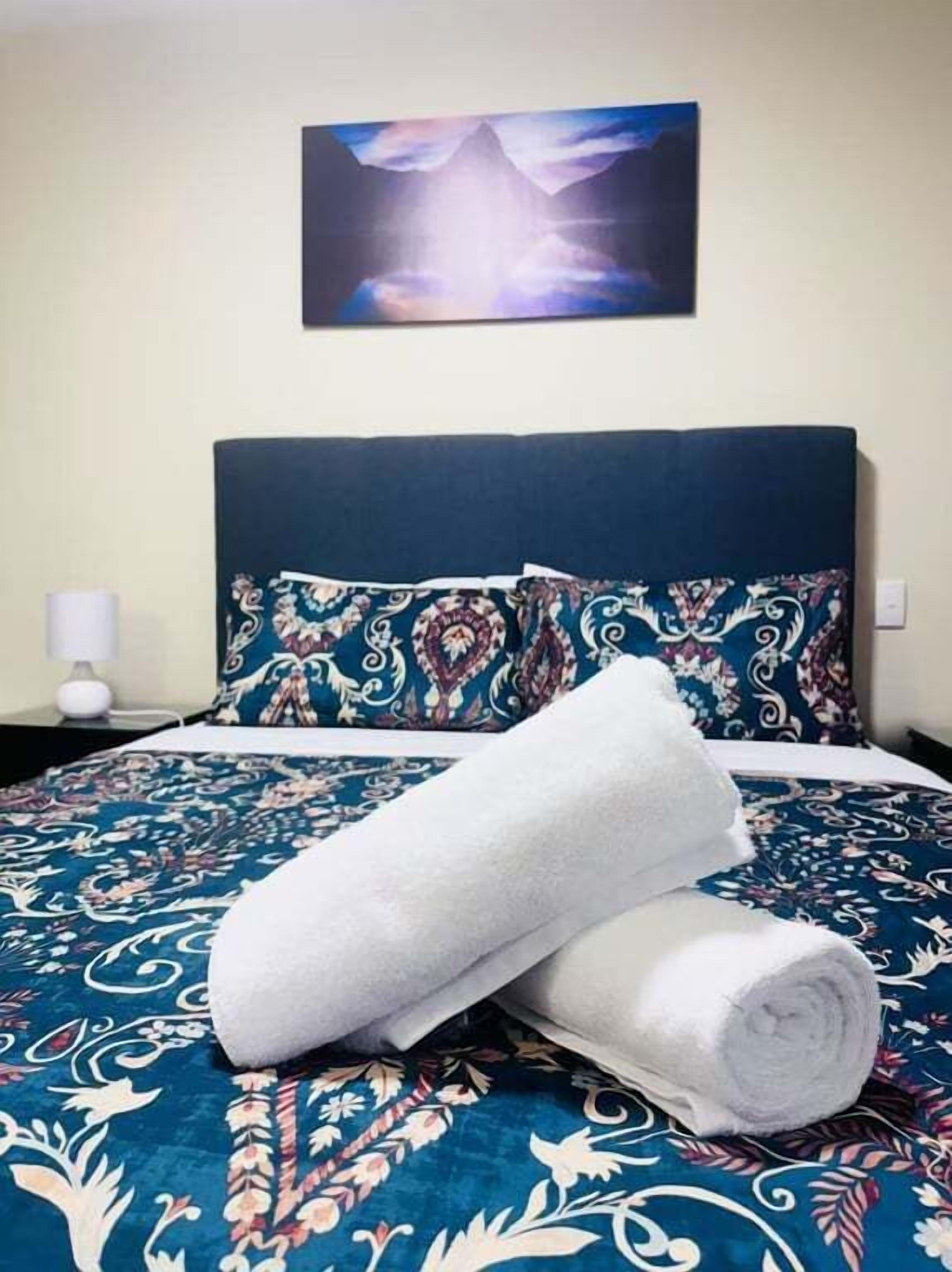 Una cama con una colcha azul y blanca estampada, dos toallas blancas enrolladas y un cuadro de un paisaje montañoso en la pared.